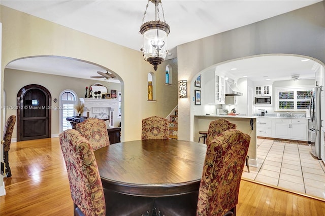 dining area featuring stairs, light wood-type flooring, a fireplace, and ceiling fan with notable chandelier