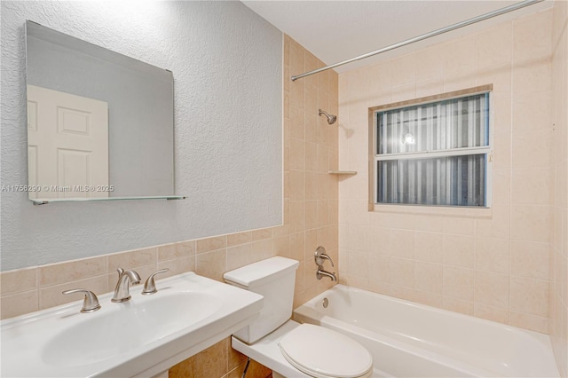 bathroom featuring bathing tub / shower combination, toilet, a textured wall, a sink, and tile walls