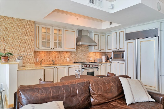 kitchen featuring built in appliances, cream cabinets, a sink, wall chimney exhaust hood, and a tray ceiling