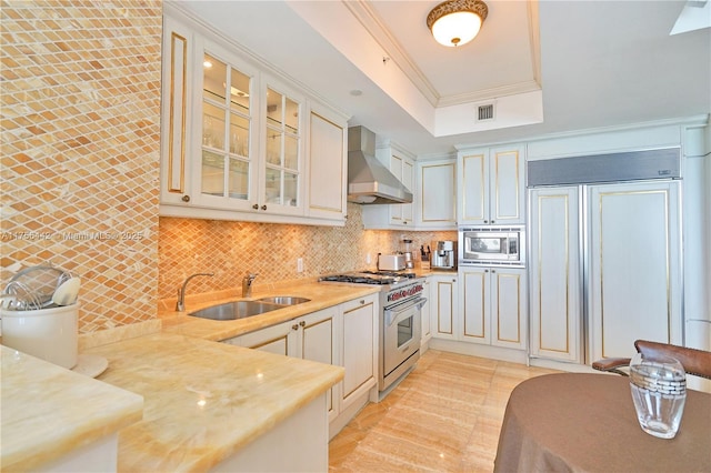 kitchen featuring wall chimney exhaust hood, ornamental molding, built in appliances, a tray ceiling, and a sink