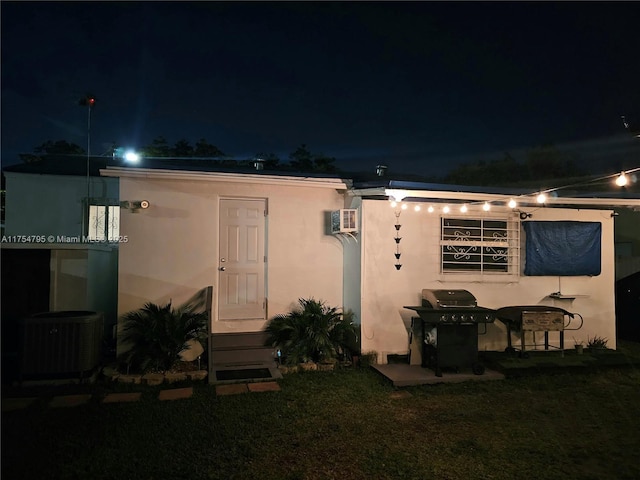 back of house at night with stucco siding, a wall unit AC, a lawn, and central air condition unit