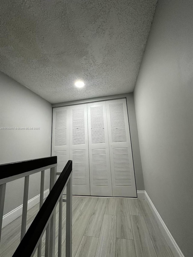 corridor featuring baseboards, a textured ceiling, an upstairs landing, and light wood-style floors