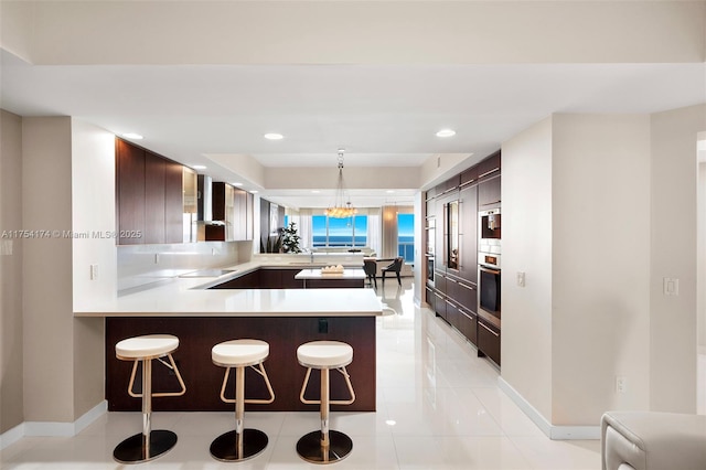 kitchen featuring a peninsula, a breakfast bar area, wall chimney exhaust hood, light countertops, and stainless steel oven