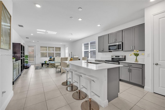 kitchen featuring light tile patterned floors, gray cabinetry, a sink, appliances with stainless steel finishes, and an island with sink