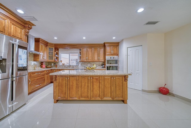 kitchen with light stone counters, stainless steel appliances, visible vents, brown cabinetry, and wall chimney range hood