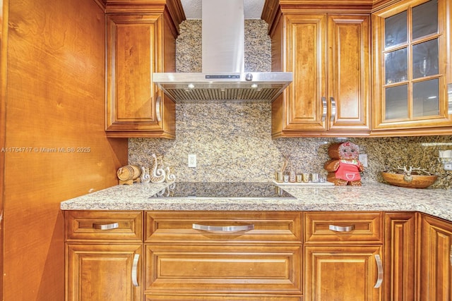 kitchen with tasteful backsplash, black electric stovetop, wall chimney range hood, glass insert cabinets, and brown cabinetry