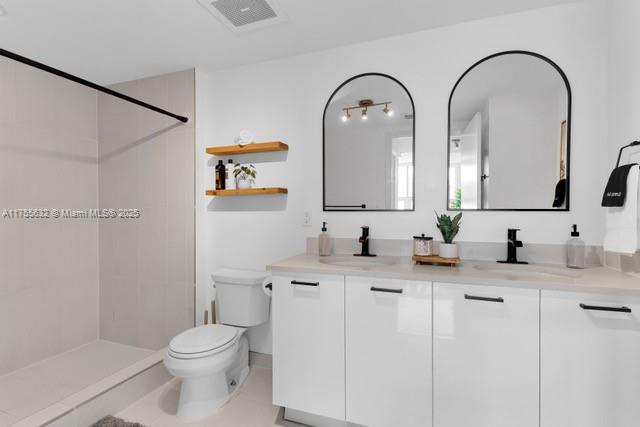 bathroom featuring double vanity, visible vents, toilet, a tile shower, and a sink
