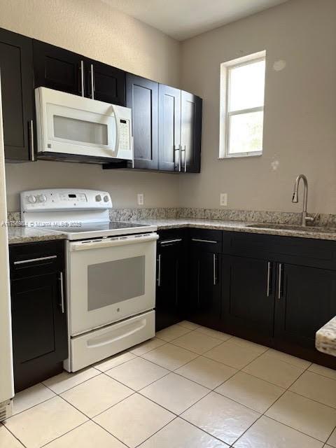 kitchen featuring light tile patterned floors, dark cabinets, white appliances, a sink, and light stone countertops