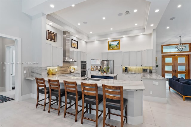 kitchen with a peninsula, a sink, a towering ceiling, appliances with stainless steel finishes, and wall chimney exhaust hood