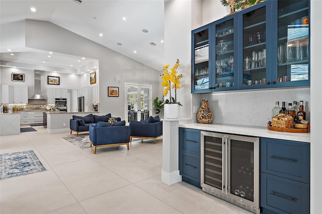 bar with light tile patterned floors, beverage cooler, a bar, wall chimney range hood, and high vaulted ceiling