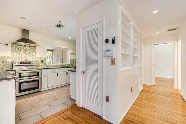 kitchen with range with two ovens, light wood-style flooring, white cabinets, wall chimney exhaust hood, and dark countertops