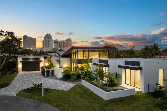 modern home featuring an attached garage, a city view, decorative driveway, a lawn, and stucco siding