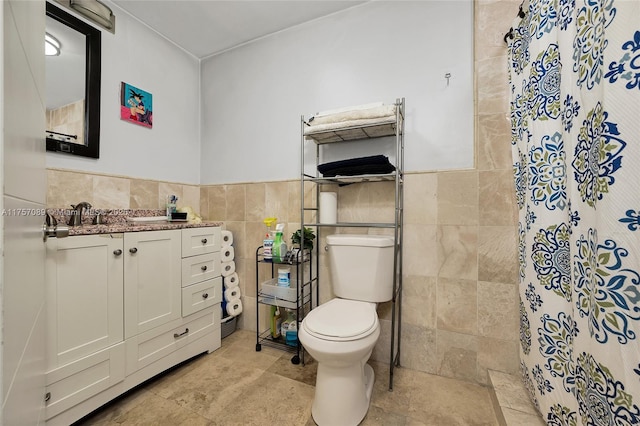 full bathroom with tiled shower, wainscoting, toilet, vanity, and tile walls