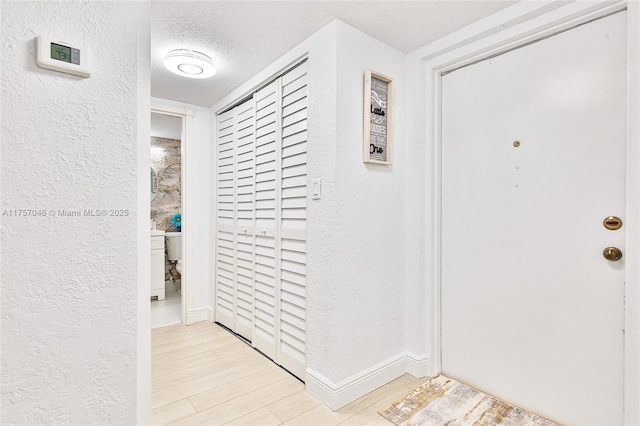 corridor featuring light wood finished floors, baseboards, a textured ceiling, and a textured wall