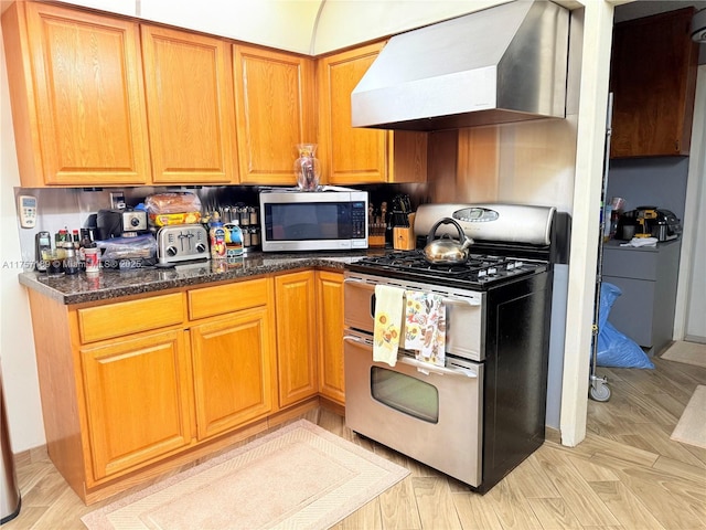 kitchen featuring brown cabinetry, appliances with stainless steel finishes, dark stone counters, and wall chimney exhaust hood