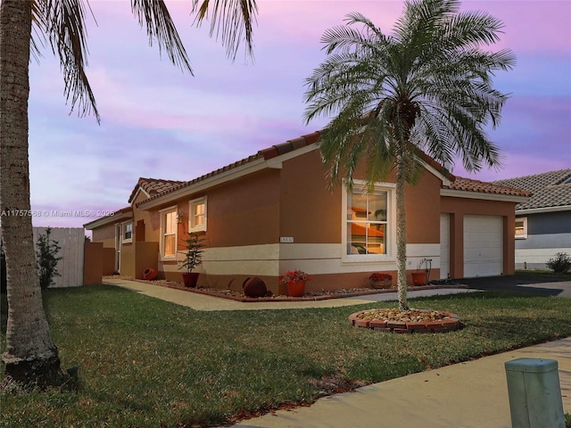 property exterior at dusk featuring a garage, a lawn, aphalt driveway, fence, and stucco siding