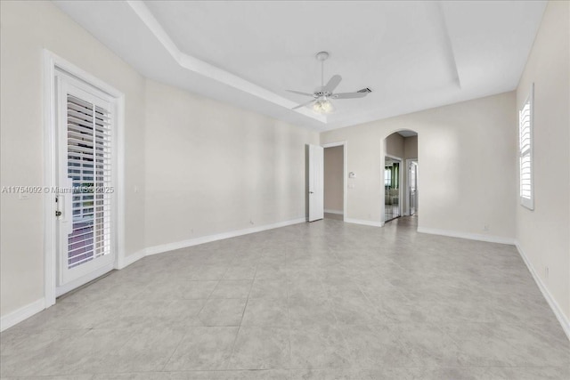 empty room featuring arched walkways, ceiling fan, a raised ceiling, and baseboards