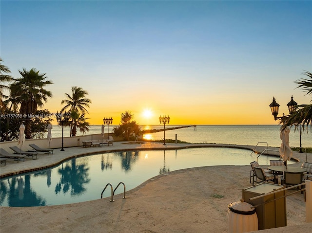 pool at dusk with a patio area, a water view, and an outdoor pool