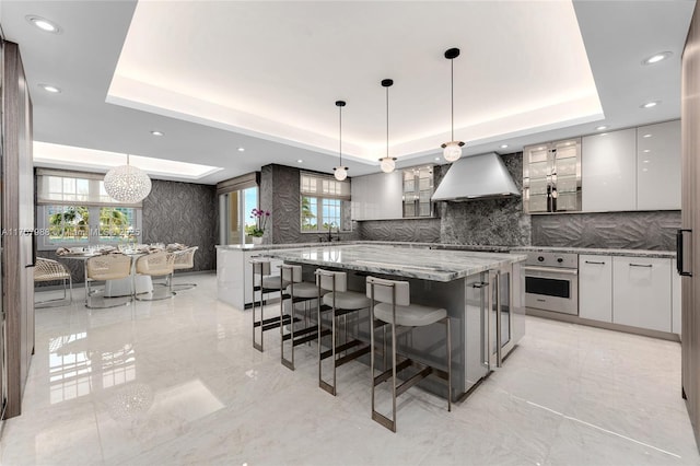 kitchen featuring a raised ceiling, stainless steel oven, wall chimney range hood, modern cabinets, and a kitchen breakfast bar