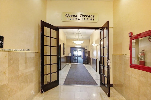 hallway with a wainscoted wall, tile patterned floors, tile walls, and french doors