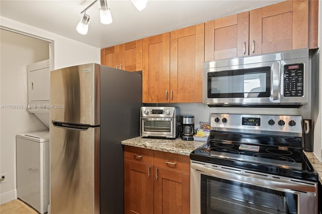 kitchen with stainless steel appliances, brown cabinetry, stacked washing maching and dryer, and light stone countertops