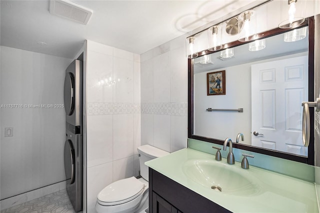 bathroom with tile walls, stacked washer and dryer, visible vents, toilet, and vanity