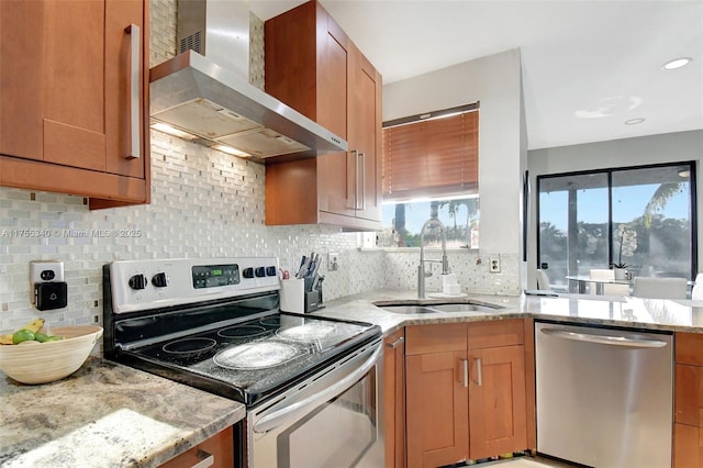 kitchen with wall chimney exhaust hood, brown cabinets, light stone countertops, stainless steel appliances, and a sink