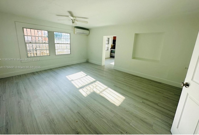 unfurnished bedroom featuring a wall unit AC, a ceiling fan, baseboards, and wood finished floors