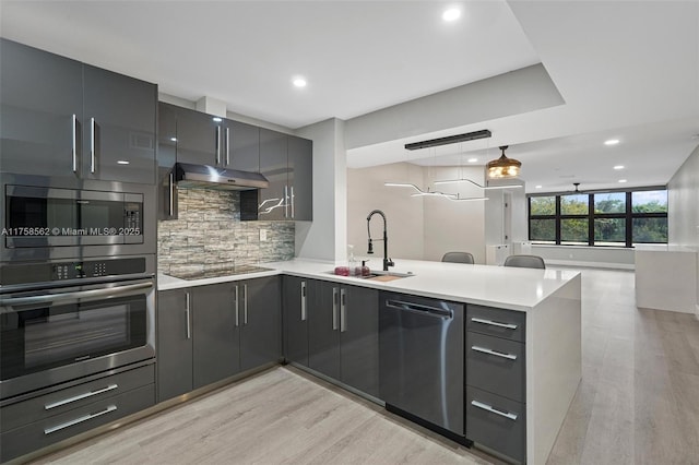 kitchen with under cabinet range hood, stainless steel appliances, a peninsula, light wood finished floors, and tasteful backsplash