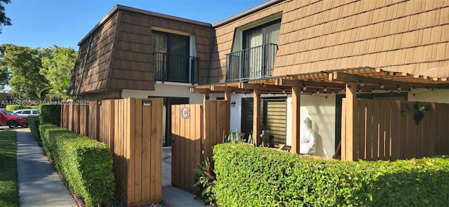 view of home's exterior with mansard roof, fence, stucco siding, and a pergola