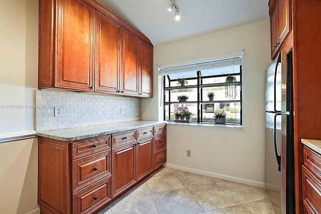 kitchen with decorative backsplash, light stone counters, brown cabinets, freestanding refrigerator, and a textured ceiling