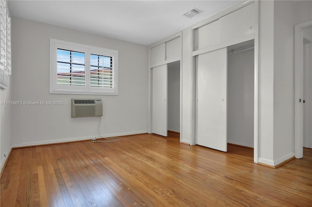 unfurnished bedroom featuring a wall unit AC, two closets, visible vents, light wood-type flooring, and baseboards