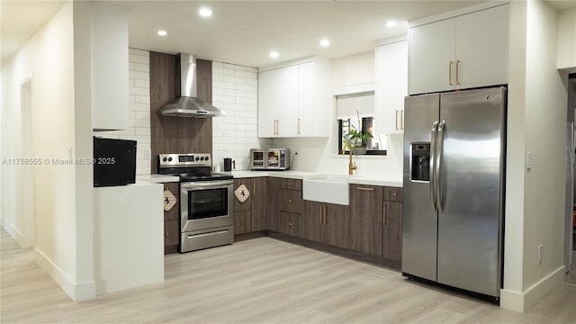 kitchen featuring tasteful backsplash, wall chimney exhaust hood, appliances with stainless steel finishes, light countertops, and light wood-style floors