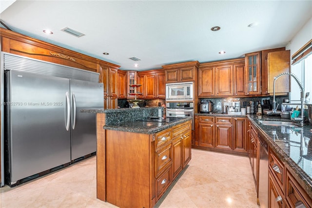 kitchen featuring brown cabinets, a kitchen island, a sink, dark stone countertops, and built in appliances