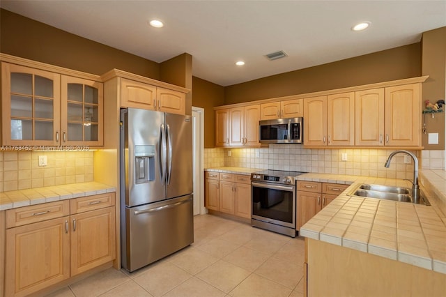 kitchen featuring light tile patterned floors, light brown cabinetry, a sink, glass insert cabinets, and appliances with stainless steel finishes