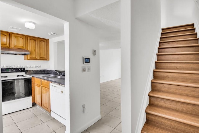 kitchen with dark countertops, light tile patterned flooring, a sink, white appliances, and under cabinet range hood