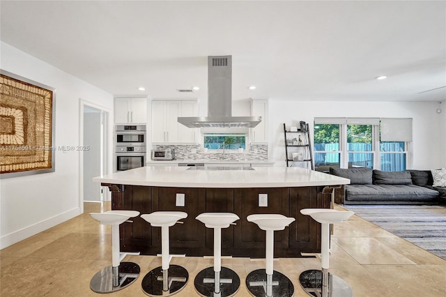 kitchen with island exhaust hood, a large island, decorative backsplash, double oven, and white cabinetry