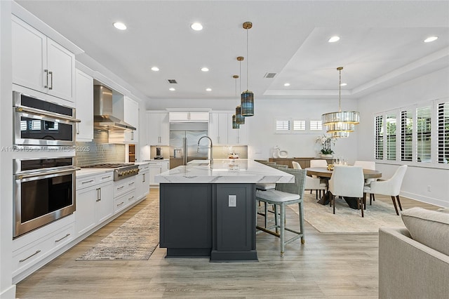 kitchen featuring wall chimney exhaust hood, a center island with sink, white cabinetry, and stainless steel appliances