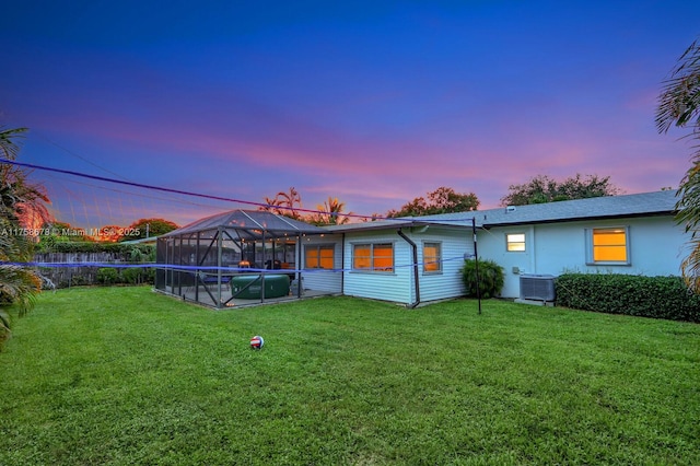 back of property at dusk featuring glass enclosure, a swimming pool, a lawn, and central AC