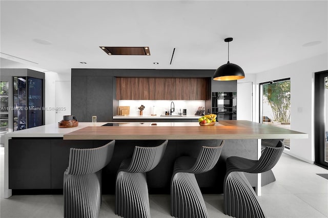 kitchen featuring dobule oven black, light countertops, hanging light fixtures, modern cabinets, and a kitchen bar