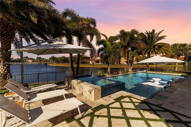 pool at dusk with a patio area, a pool with connected hot tub, a water view, and fence