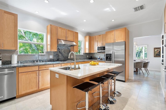 kitchen featuring light stone counters, appliances with stainless steel finishes, under cabinet range hood, a kitchen island with sink, and visible vents