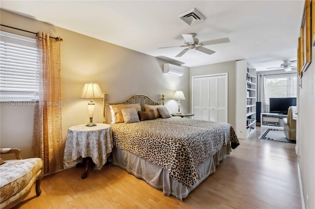 bedroom with a wall unit AC, light wood finished floors, a closet, visible vents, and a ceiling fan