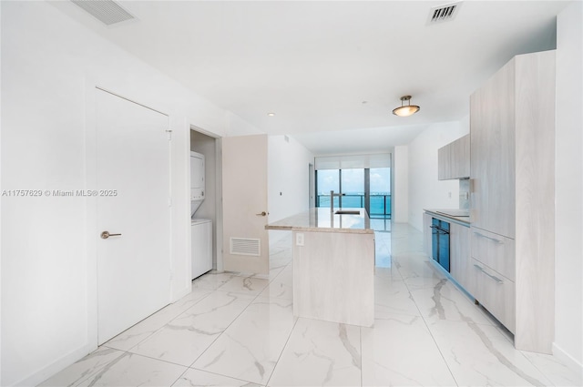 kitchen featuring marble finish floor, stacked washer and dryer, modern cabinets, and a kitchen island with sink