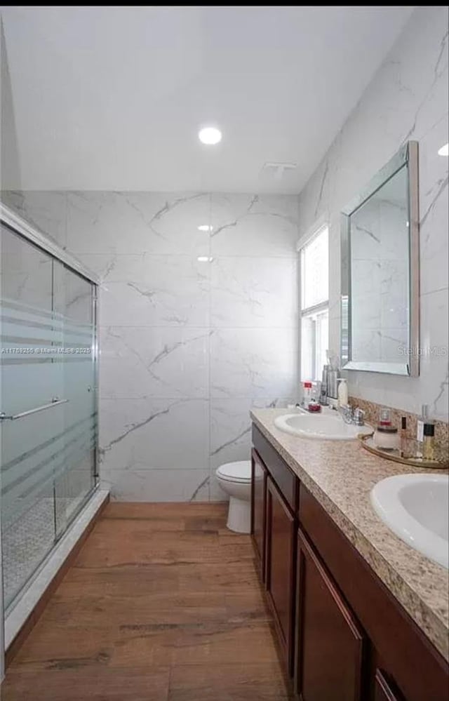 bathroom featuring toilet, wood finished floors, a sink, a marble finish shower, and double vanity