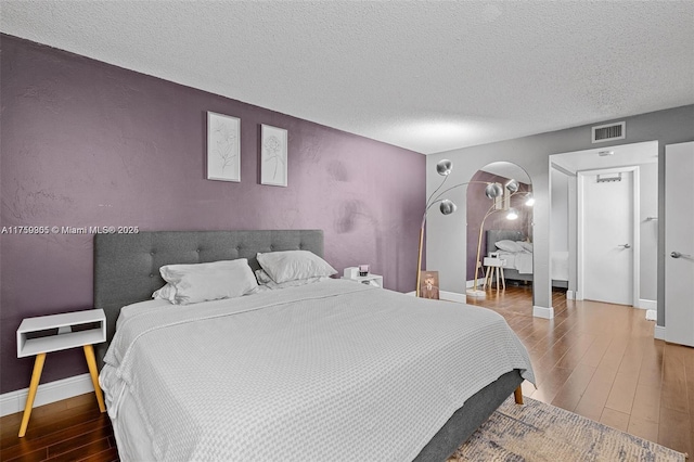 bedroom with a textured ceiling, arched walkways, wood finished floors, visible vents, and baseboards