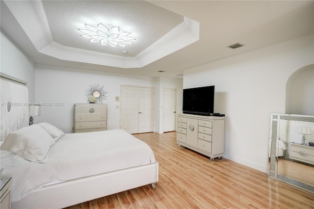 bedroom with arched walkways, light wood finished floors, a raised ceiling, visible vents, and baseboards