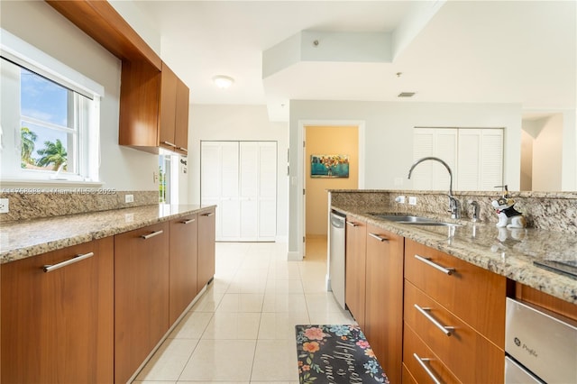 kitchen featuring light tile patterned floors, a sink, stainless steel dishwasher, light stone countertops, and brown cabinetry