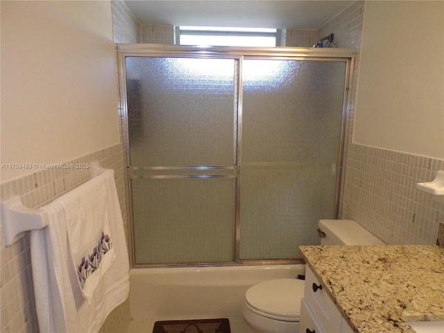 full bath with toilet, a wainscoted wall, combined bath / shower with glass door, vanity, and tile walls