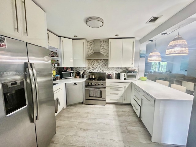 kitchen featuring stainless steel appliances, light countertops, decorative backsplash, a peninsula, and wall chimney exhaust hood